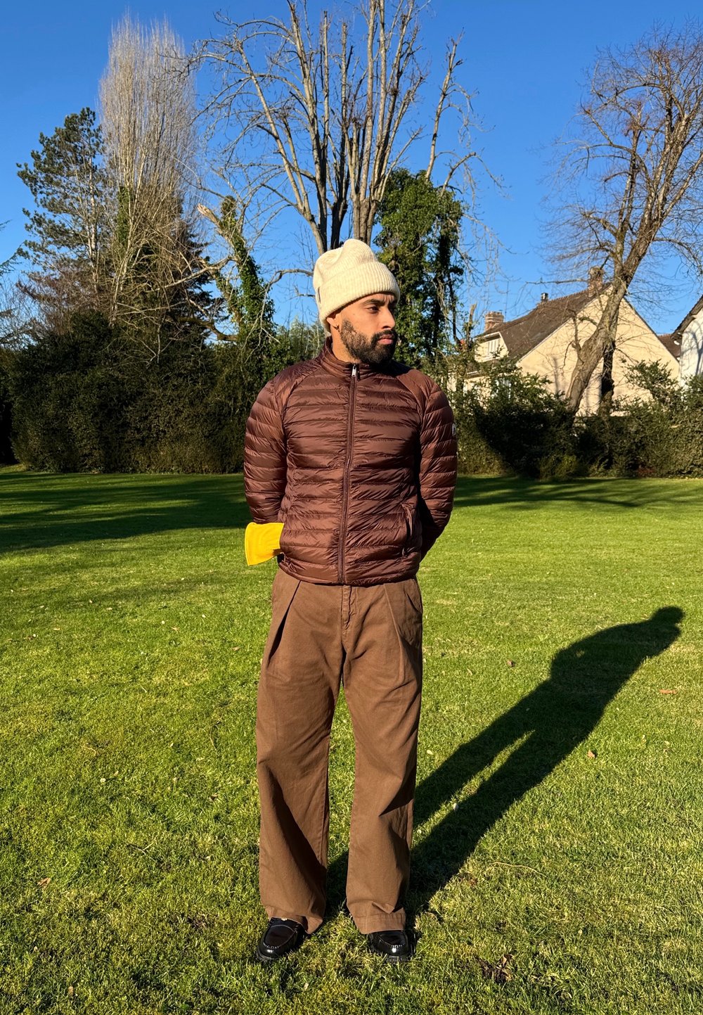 Braune Puffjacke mit hohem Kragen, beiger Strickmütze, lockere braune Hose und schwarze Schuhe, stehend auf grünem Gras mit Bäumen im Hintergrund.
