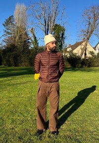 Braune Puffjacke mit hohem Kragen, beiger Strickmütze, lockere braune Hose und schwarze Schuhe, stehend auf grünem Gras mit Bäumen im Hintergrund.