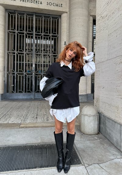 Camiseta negra de gran tamaño superpuesta a una camisa blanca de botones, con botas negras hasta la rodilla y un bolso negro. Entorno urbano con paredes texturizadas.