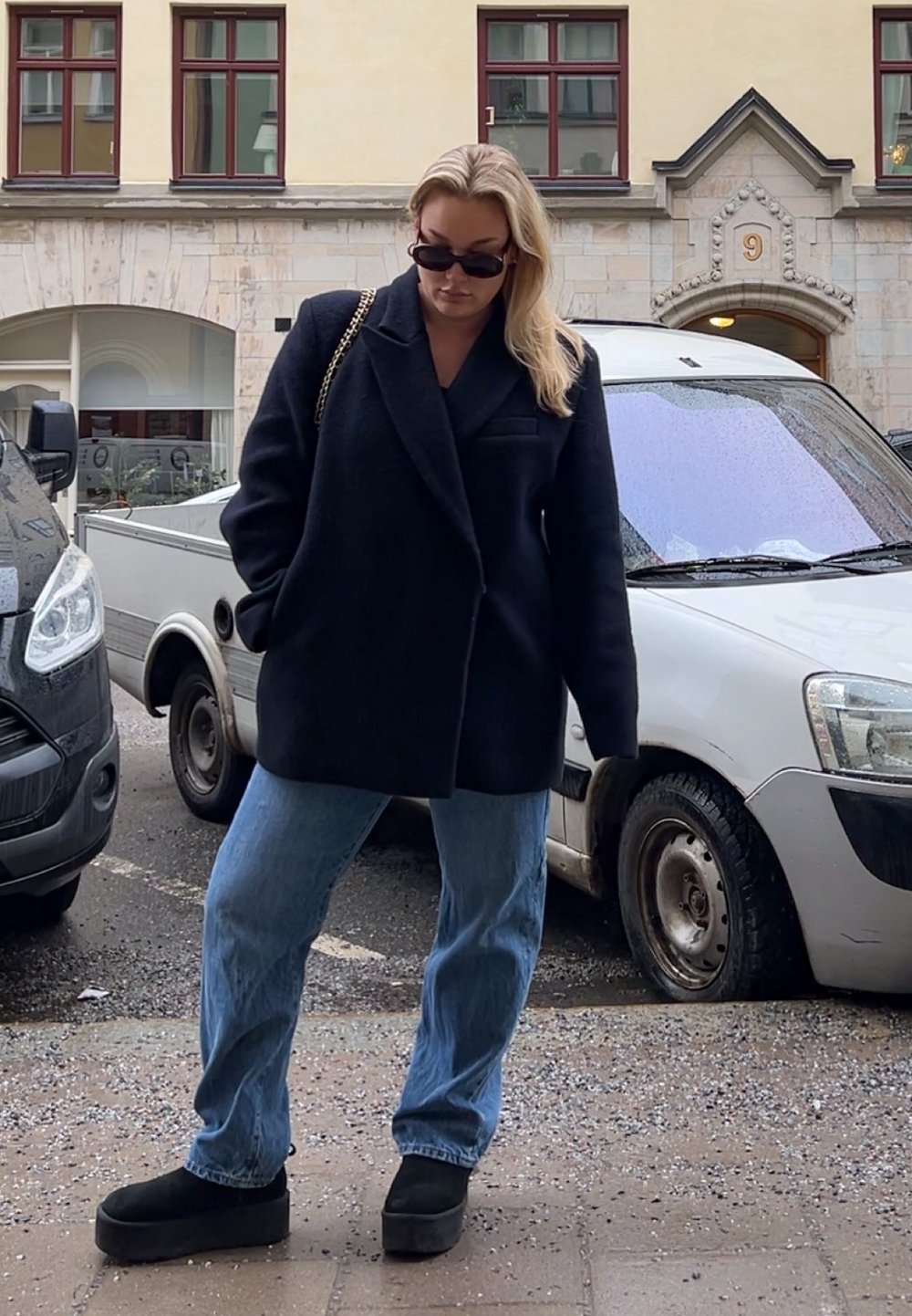 Schwarzer Wollmischungsmantel mit übergroßen Revers, hellblaue gerade Jeans und schwarze Plateaustiefel. Schwarze Sonnenbrille und Tasche mit Kettenakzenten.