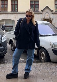 Schwarzer Wollmischungsmantel mit übergroßen Revers, hellblaue gerade Jeans und schwarze Plateaustiefel. Schwarze Sonnenbrille und Tasche mit Kettenakzenten.