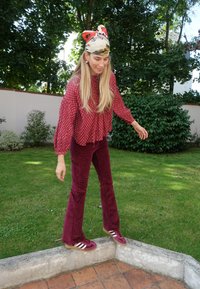 Burgunder Cord-Hosen mit weit geschnittenen Beinen, kombiniert mit einer gemusterten kastanienbraunen Bluse. Das Model trägt ein buntes Kopftuch und Sneakers mit weißen Streifen.