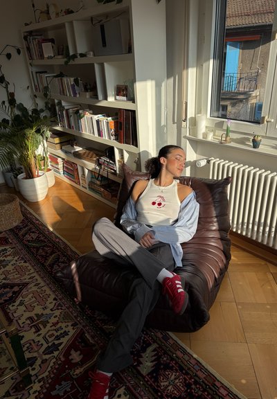 Una silla de frijoles de cuero marrón oscuro sostiene a una persona sentada, que lleva una camiseta sin mangas blanca con cerezas rojas, pantalones grises y zapatillas rojas.