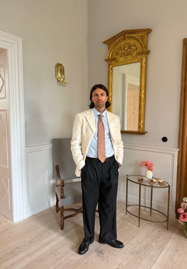 Homme en blazer blanc, chemise bleue, cravate orange et pantalon noir debout dans une pièce élégante à côté d'un miroir, d'une chaise et d'une petite table avec des fleurs.
