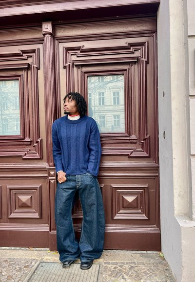 Hombre con rastas apoyado contra una puerta de madera ornamentada, lleva un suéter azul y jeans de piernas anchas, mirando hacia la izquierda.