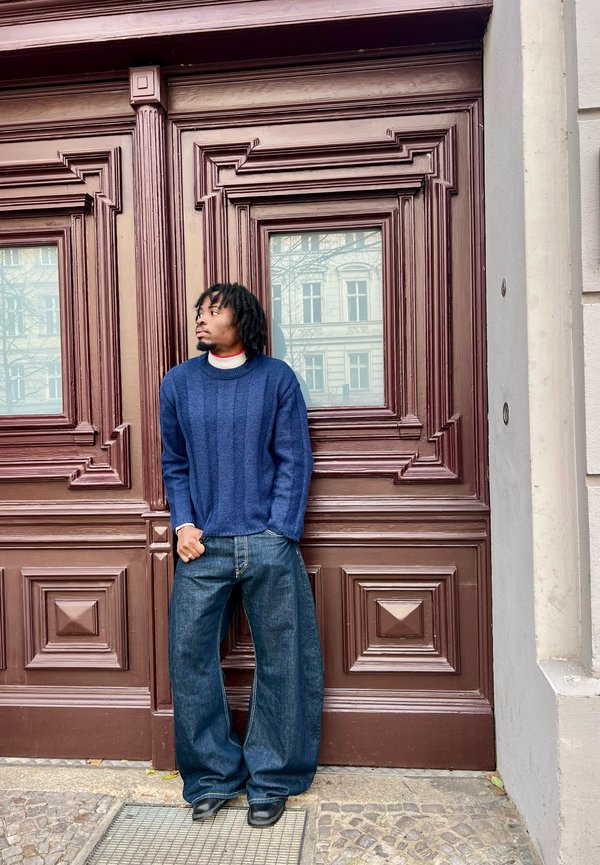 Mann mit Dreadlocks steht vor einer kunstvoll verzierten Holztür, trägt einen blauen Pullover und eine weit geschnittene Jeans und blickt nach links.