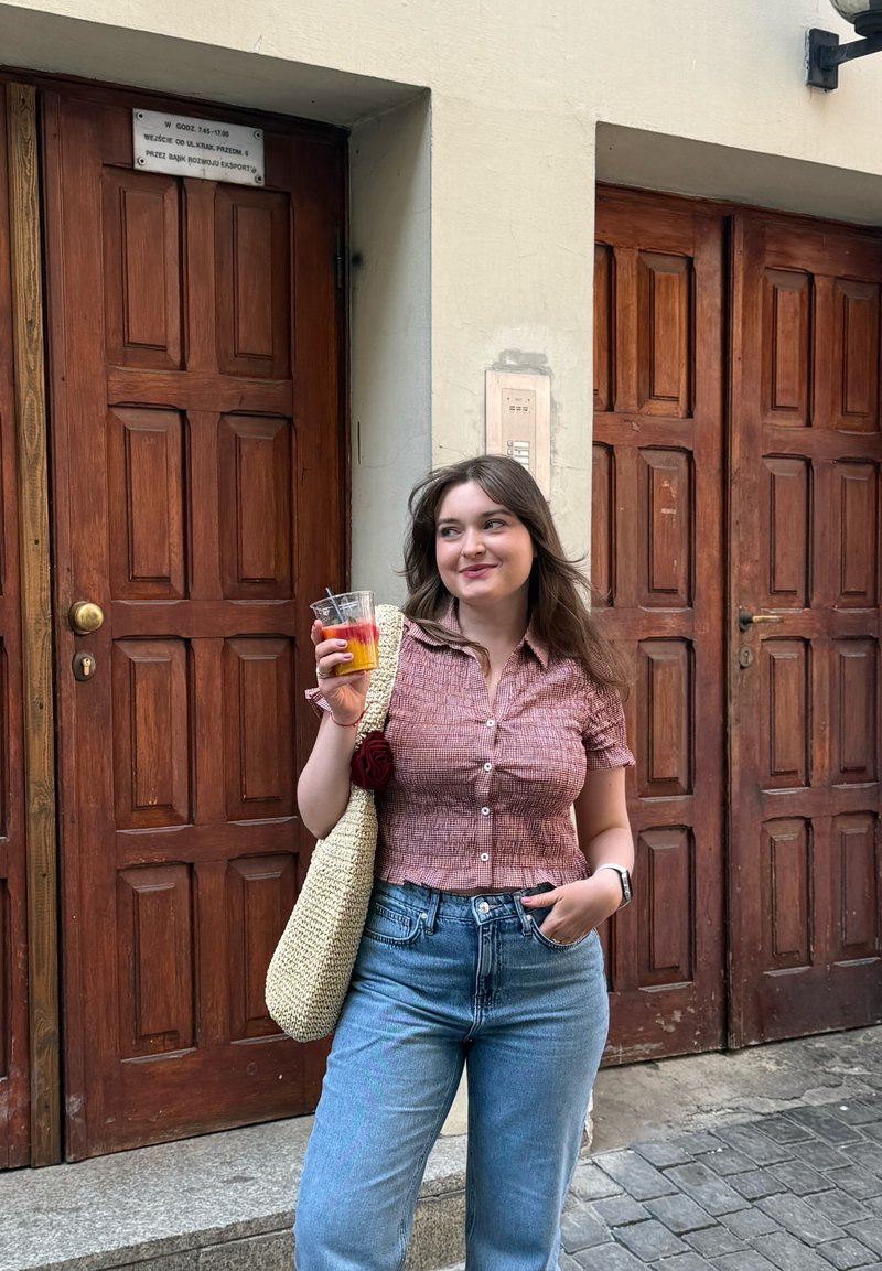 Jeune femme tenant une boisson aux fruits en couches, portant une chemise à carreaux rouge et un jean, debout devant des portes en bois dans une rue de la ville.