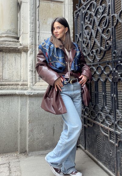 Mujer apoyada contra una elaborada puerta de metal negra, vistiendo una chaqueta de cuero marrón, bufanda azul, vaqueros azul claro y zapatillas deportivas al aire libre.