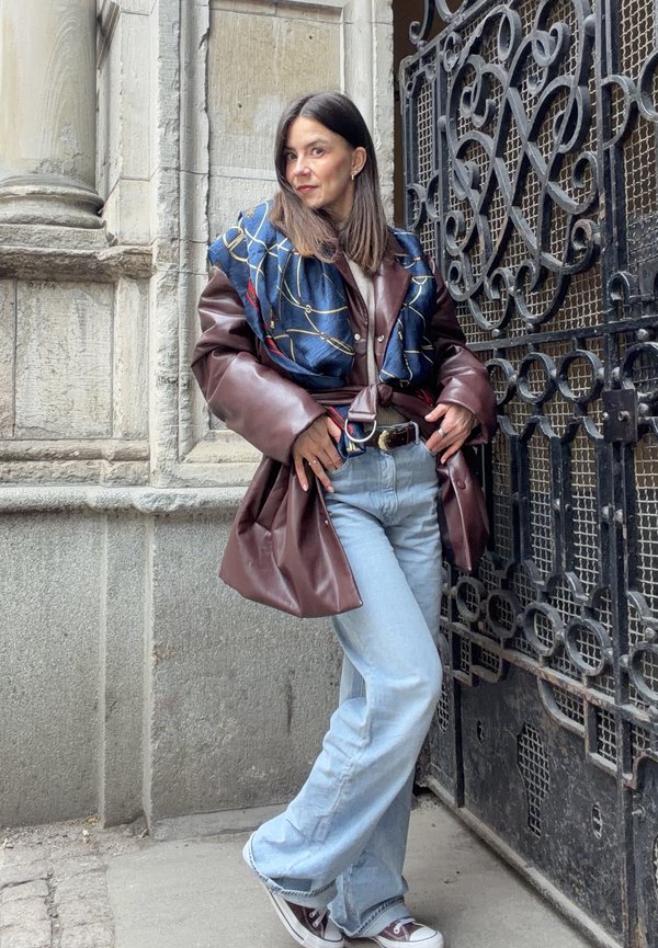 Nő egy díszes fekete fémkapunak támaszkodik, barna bőrkabátot, kék sálat, világoskék farmert és tornacipőt visel kint.