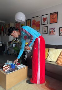 Άτομο με μπλε και κόκκινη φόρμα Adidas σκύβει πάνω από ένα ξύλινο τραπεζάκι σαλονιού με διάφορα αντικείμενα, σε ένα σαλόνι με κορνιζαρισμένη τέχνη στον τοίχο.