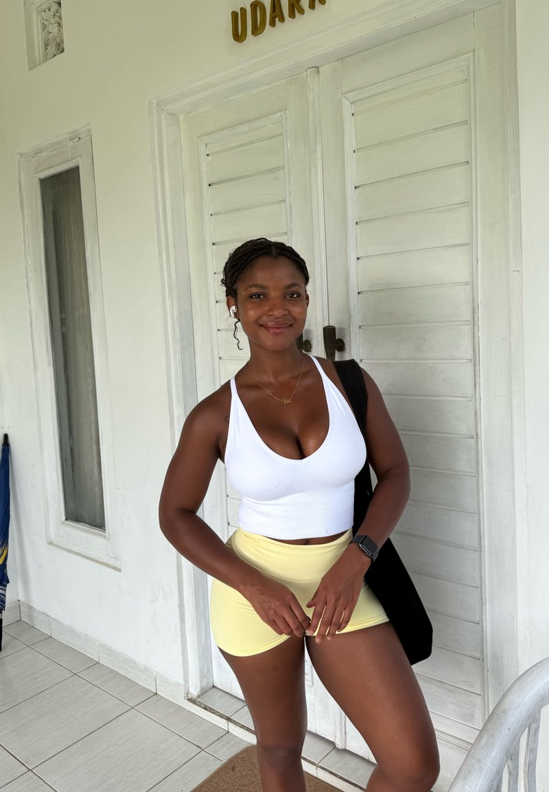 Haut court blanc et short jaune, debout devant une porte en bois blanche avec une plaque dorée. Sac noir et montre-bracelet visibles.