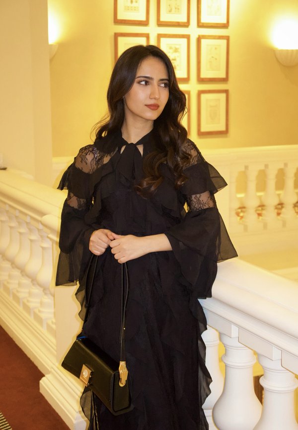 Mujer con vestido negro de encaje sosteniendo un bolso negro, de pie junto a una barandilla blanca en un interior iluminado cálidamente con arte enmarcado en la pared.