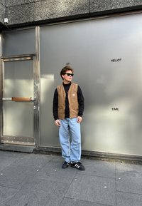 Gilet di tessuto beige sopra un dolcevita nero, jeans baggy azzurri e scarpe nere con strisce bianche. Lo sfondo presenta un muro e una porta grigi.