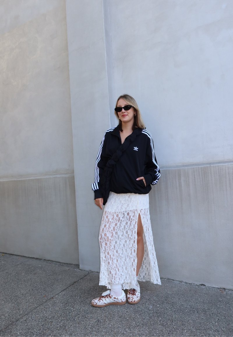 Veste de sport noire avec des rayures blanches, jupe maxi blanche en dentelle avec une fente sur le côté, chaussettes blanches, et chaussures marron à motif.