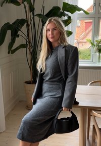 Grijze wollen blazer over een grijze wollen polo en rok, gecombineerd met een kleine zwarte handtas. Binnenomgeving met planten en een houten tafel.