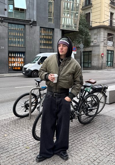 Chaqueta bomber verde oliva con cremallera y bolsillo; pantalones vaqueros negros anchos; zapatos negros; sosteniendo una taza de café; bicicletas de fondo.