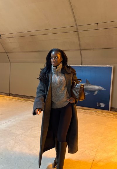 Mujer con abrigo gris, suéter, mallas negras y botas camina en una estación con un cartel de delfines en el fondo.