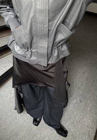 Personne portant une veste en cuir grise, une jupe sombre superposée sur un pantalon ample sombre, et des chaussures noires brillantes à bout carré, debout à l'intérieur.