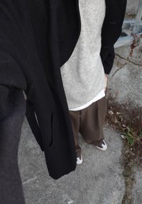Cappotto di lana nero con grandi rever, indossato sopra un maglione grigio chiaro e una camicia bianca. Pantaloni marroni a righe sottili e sneakers completano il look.