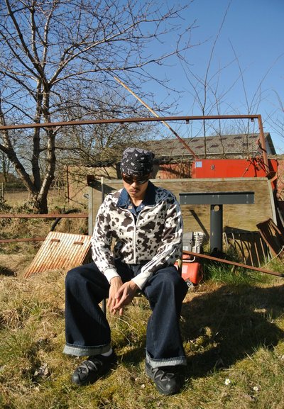 Chaqueta con cremallera con estampado de vaca, puños y cuello blancos. Jeans de pierna ancha de color azul oscuro. Calzado negro. Entorno al aire libre con césped y árboles.