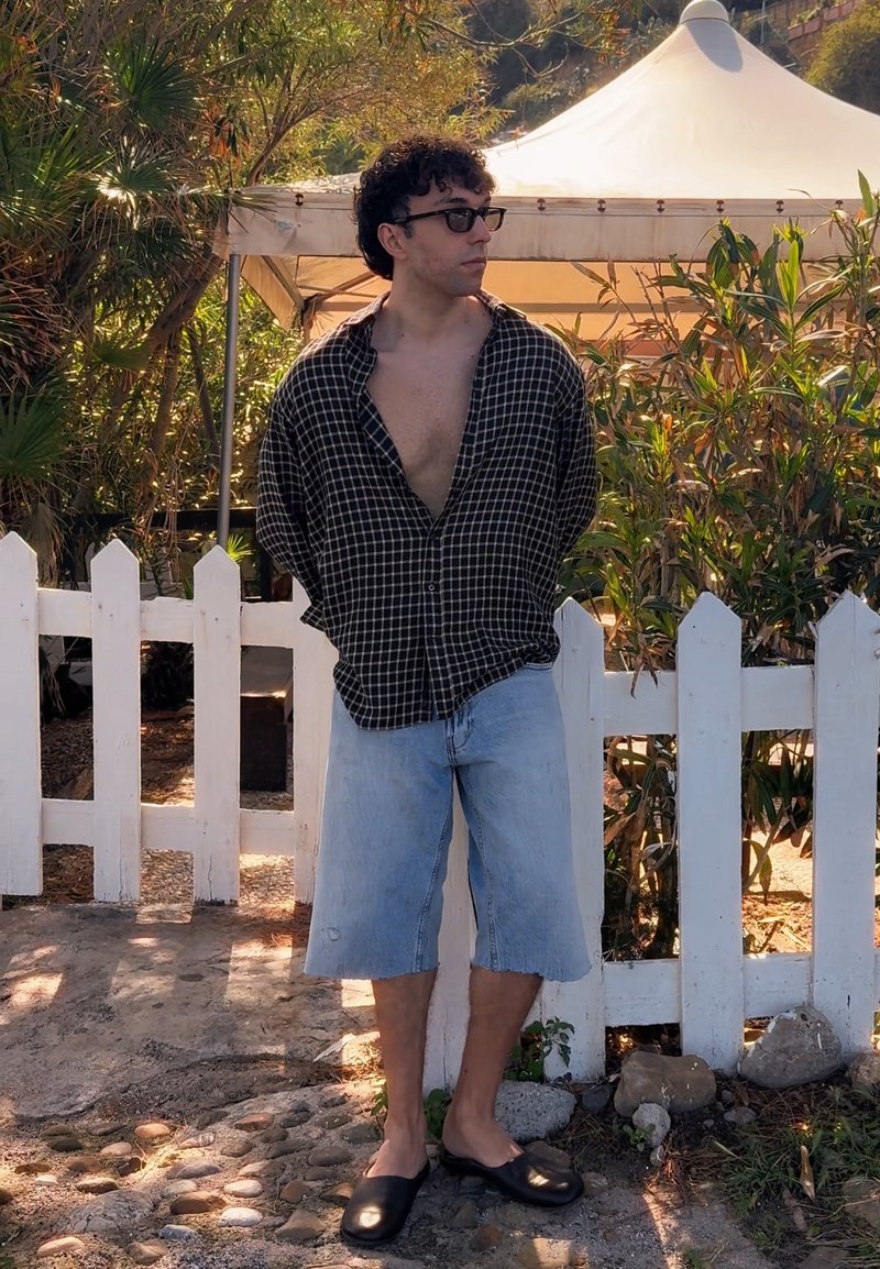 Homme aux cheveux bouclés et lunettes de soleil debout près d'une clôture blanche en portant une chemise ouverte, un short en jean et des chaussures noires sans lacets en plein air.