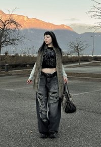 Zwart cropped top met schedelontwerp, grijze gescheurde wijde pijpen spijkerbroek, bontvest, zwarte riem en een handtas vasthoudend, tegen een bergachtergrond.