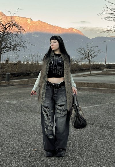 Top corto negro con diseño de calavera, jeans anchos gris desgarrados, chaleco de piel, cinturón negro y bolso en mano, con un paisaje montañoso de fondo.