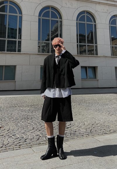 Persona con cabello rosa que lleva una chaqueta negra oversize, camisa blanca, pantalones cortos negros y botas negras, de pie en una calle empedrada junto a un edificio con ventanas.