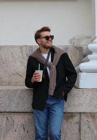 Sort dobbeltradet frakke, beige cardigan draperet om skuldrene, hvid t-shirt, blå jeans, holder en iskold drik med turkise sugerør.