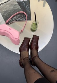 Zapatos de tacón cuadrado de cuero marrón con punta cuadrada y correa en el tobillo, combinados con medias negras transparentes. Bolso rosa y bebida verde sobre una mesa.