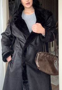 Haină neagră de blană artificială cu guler pufos, design cu două rânduri de nasturi și mâneci largi, asortată cu o geantă mică de mână din piele maro.