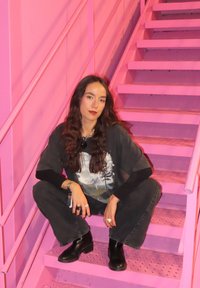 Čierne kožené členkové topánky, veľké čierne tričko s potlačou hôr, tmavé nohavice, dlhé vlnité vlasy na pozadí ružového schodiska.