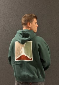 Grønn hettegenser med en stor Jordan-logo på baksiden. Mykt stoff, oversized passform, ribbestrikkede mansjetter og nederkant. Strukturert mørk bakgrunn.