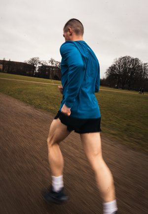 Hombre con camisa azul de manga larga y pantalones cortos negros corriendo por un camino en un parque con césped y árboles al fondo en un día nublado.