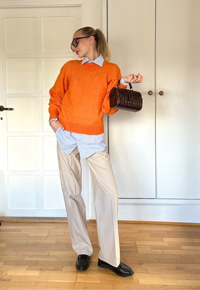 Suéter naranja de gran tamaño, camisa de rayas azules debajo, pantalones beige con raya diplomática y un pequeño bolso marrón con estampado de cocodrilo. Zapatos negros.