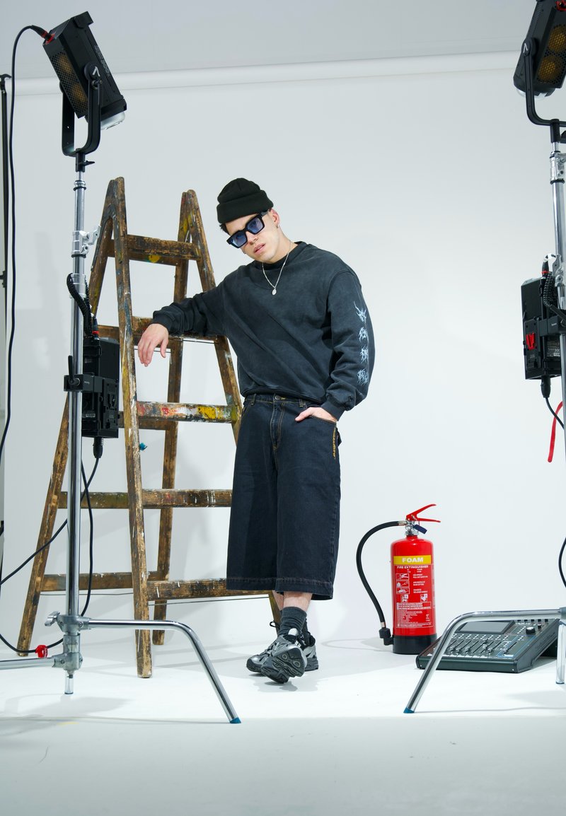Schwarzer übergroßer Sweatshirt mit Grafik, dunkelblaue Denim-Culottes und graue Sneakers. Im Hintergrund sind eine Holzleiter und ein Feuerlöscher zu sehen.