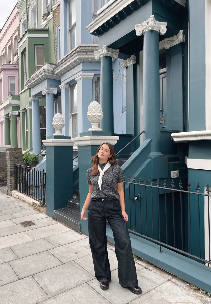 Calças pretas de perna larga com uma blusa cinza texturizada e um lenço branco, em pé em frente a casas coloridas com detalhes em colunas brancas e vasos decorativos.