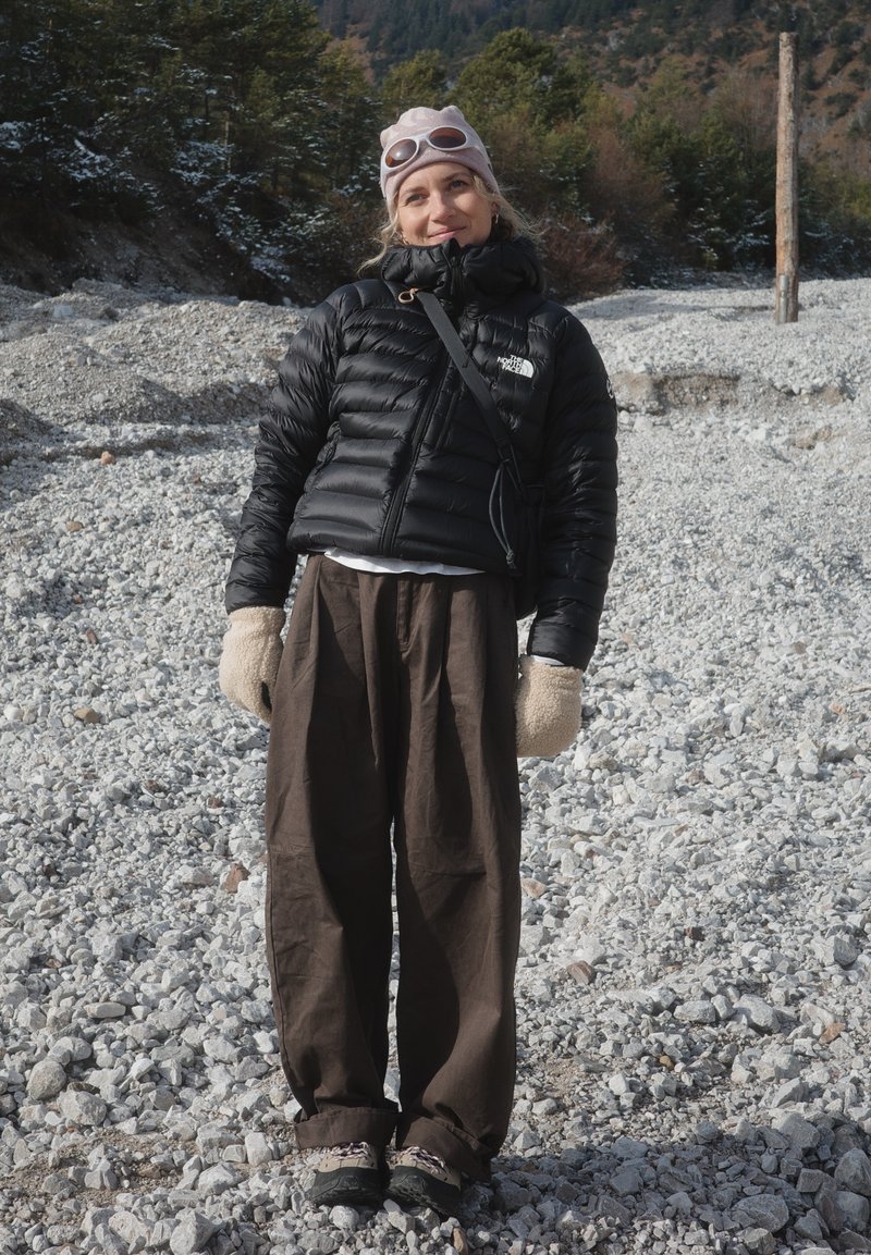Woman wearing a black puffer jacket, pink beanie, beige mittens, and brown pants standing on a rocky terrain with pine trees in the background.