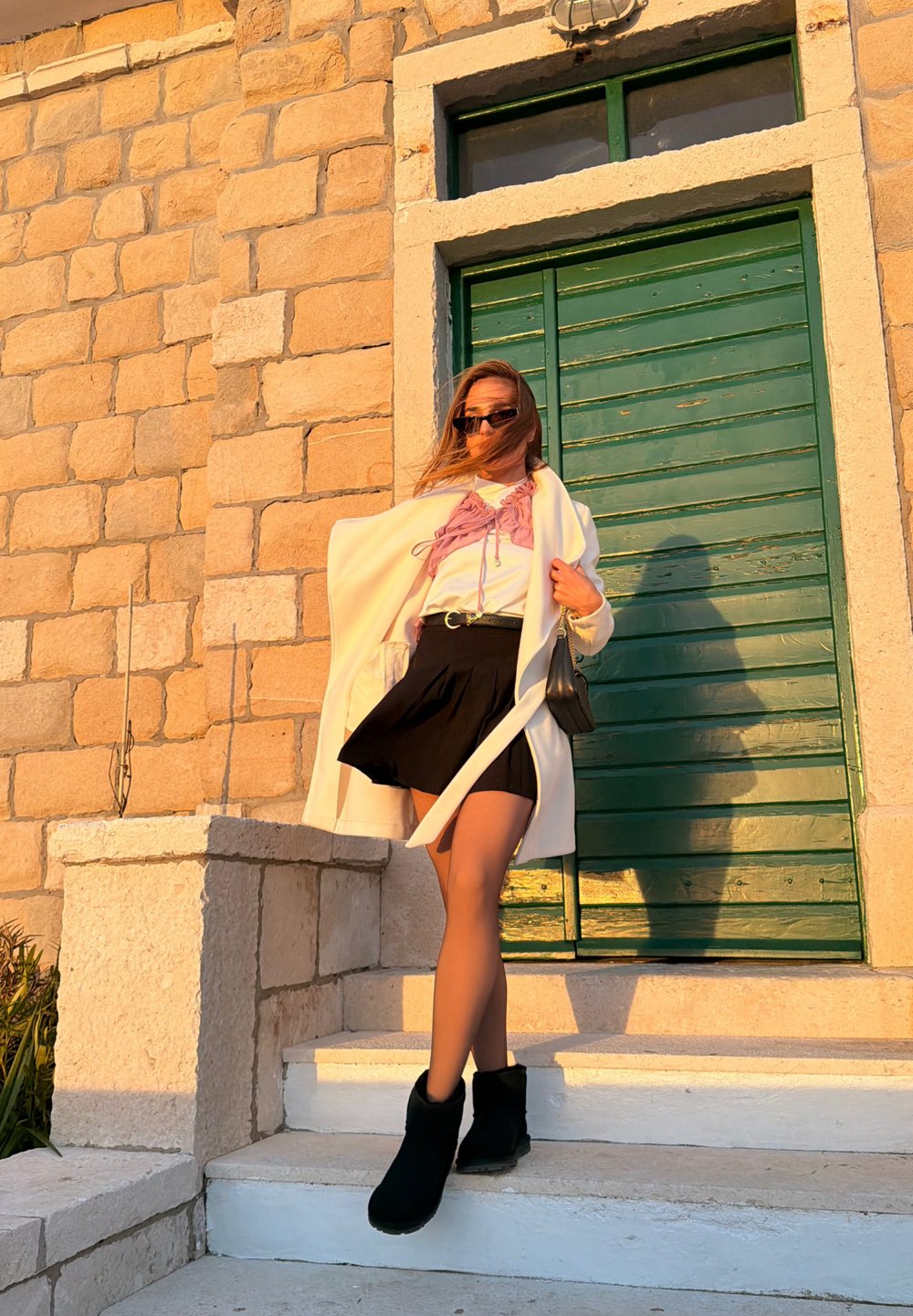 Γυναίκα με γυαλιά ηλίου, φορώντας λευκό παλτό, μαύρη φούστα και μπότες, στέκεται σε πέτρινα σκαλοπάτια κοντά σε πράσινη ξύλινη πόρτα στο ηλιοβασίλεμα.