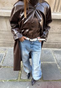 Personne portant un manteau en cuir brun foncé, une chemise à carreaux marron, un jean bleu clair avec ceinture blanche et des chaussures pointues noires, debout sur un pavé en pierre.