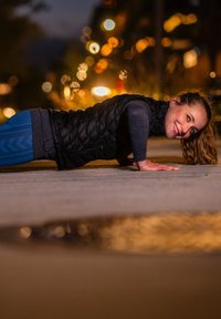 Fekete puffos mellény sötét hosszú ujjú felső és mintás kék leggings felett. A személy éjszaka fekvő támasz helyzetben végzi el a fekvőtámaszt egy járdán.