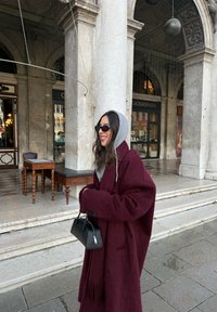 Oversized kåpe i burgunder med teksturert finish, grå hette og svart veske. Har store knapper og frynsete kant. Bakgrunn med steinlagt belegning.