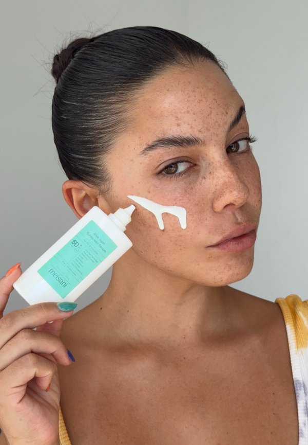 Kvinde med mørkt hår i knold påfører hvid solcreme-serum på sin fregnede kind fra en hvid flaske mærket SPF 50+ med turkise detaljer.