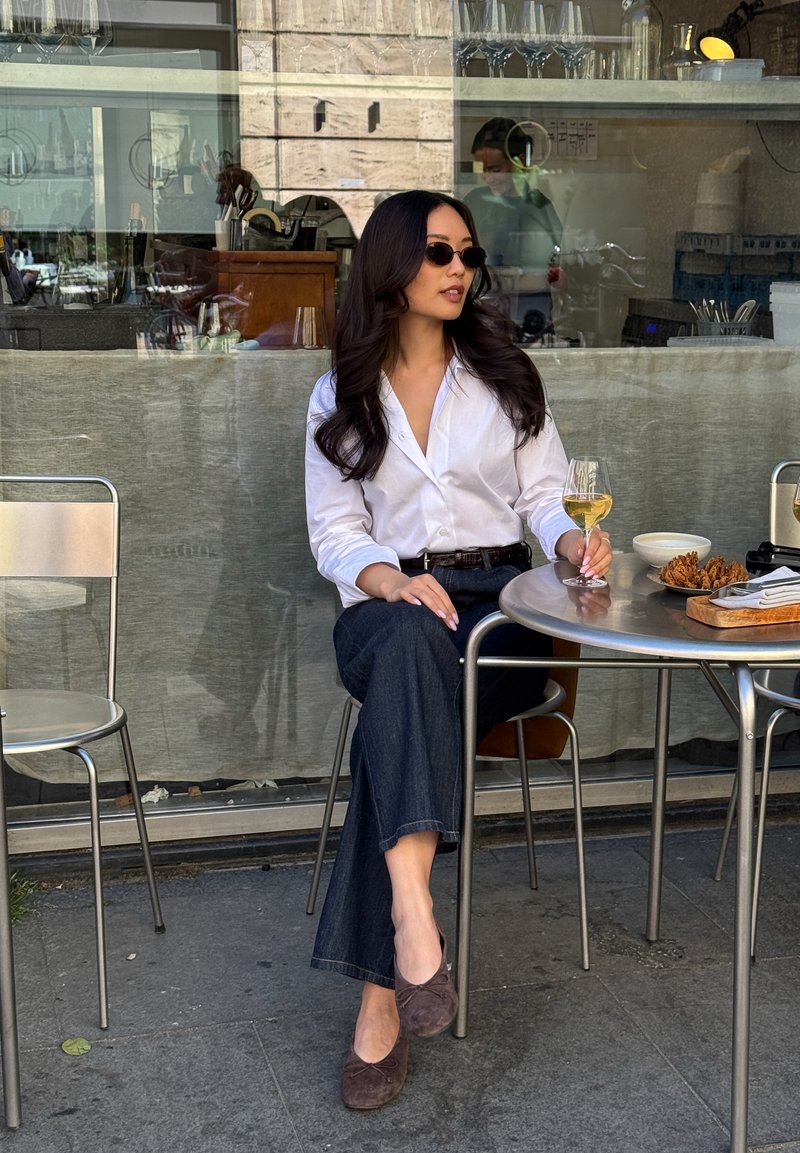 Femme en lunettes de soleil et chemise blanche assise à la table d’un café en terrasse, tenant un verre de vin, avec de la nourriture et des serviettes sur la table.