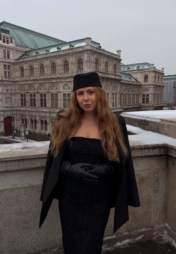 Femme aux cheveux longs et ondulés portant une robe noire, des gants, un manteau et un chapeau, debout sur une terrasse en pierre devant un bâtiment historique orné par une journée nuageuse.