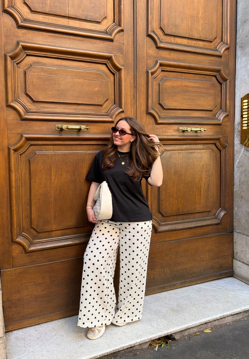 Femme en t-shirt noir, pantalon blanc à pois et lunettes de soleil, debout près de grandes portes en bois, tenant un sac blanc et souriant.