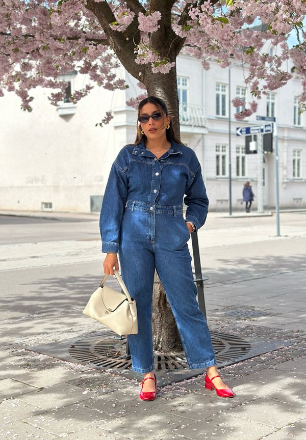 Vrouw in een spijkerjumpsuit en rode schoenen staat onder een bloeiende kersenboom op een stadsstoep en houdt een lichtbeige handtas vast.