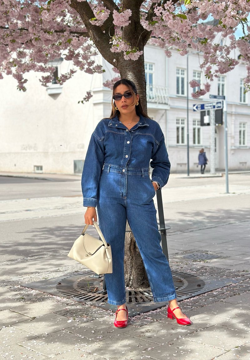 Femme portant une combinaison en jean et des chaussures rouges, debout sous un cerisier en fleurs sur un trottoir de la ville, tenant un sac à main beige clair.