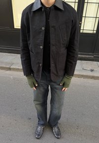 Άτομο που φοράει μαύρο σακάκι με κουμπιά και δερμάτινο γιακά, μαύρο πουκάμισο, ξεθωριασμένο μαύρο τζιν, ελαιοπράσινα γάντια χωρίς δάχτυλα και πλεκτά μαύρα παπούτσια.