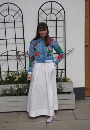 Mujer junto a una maceta con narcisos, lleva un cárdigan azul con estampado, una falda larga blanca y zapatos azul claro, delante de una pared blanca con enrejados metálicos.